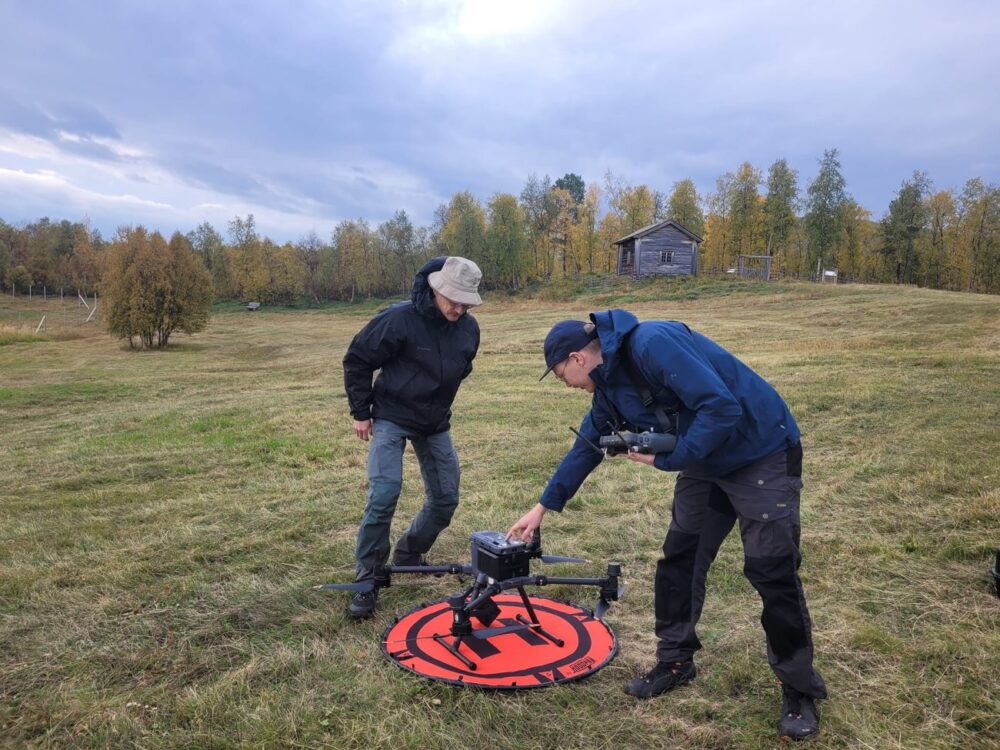 kaksi ihmistä aukiolla ja drone maassa lähtövalmiina